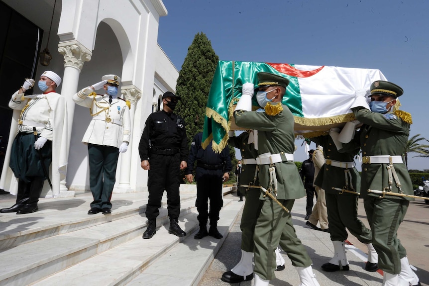 Soldiers in dress uniform carry a coffin draped in a flag past other military personnel into a white building.