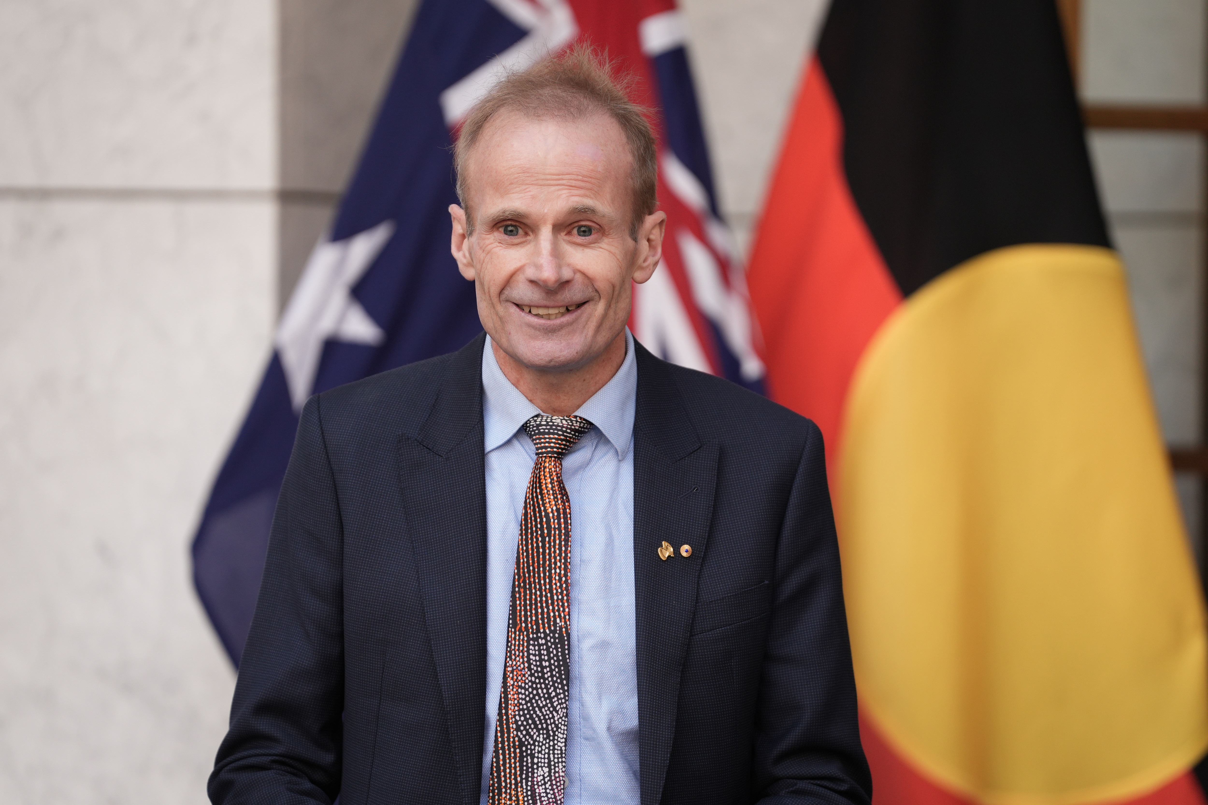 A man in a suit a tie smiles, in front of Australian and Aboriginal flags.