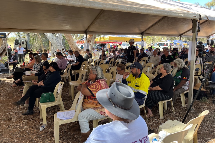 A crowd of people sitting under a marquee at a bush meeting.