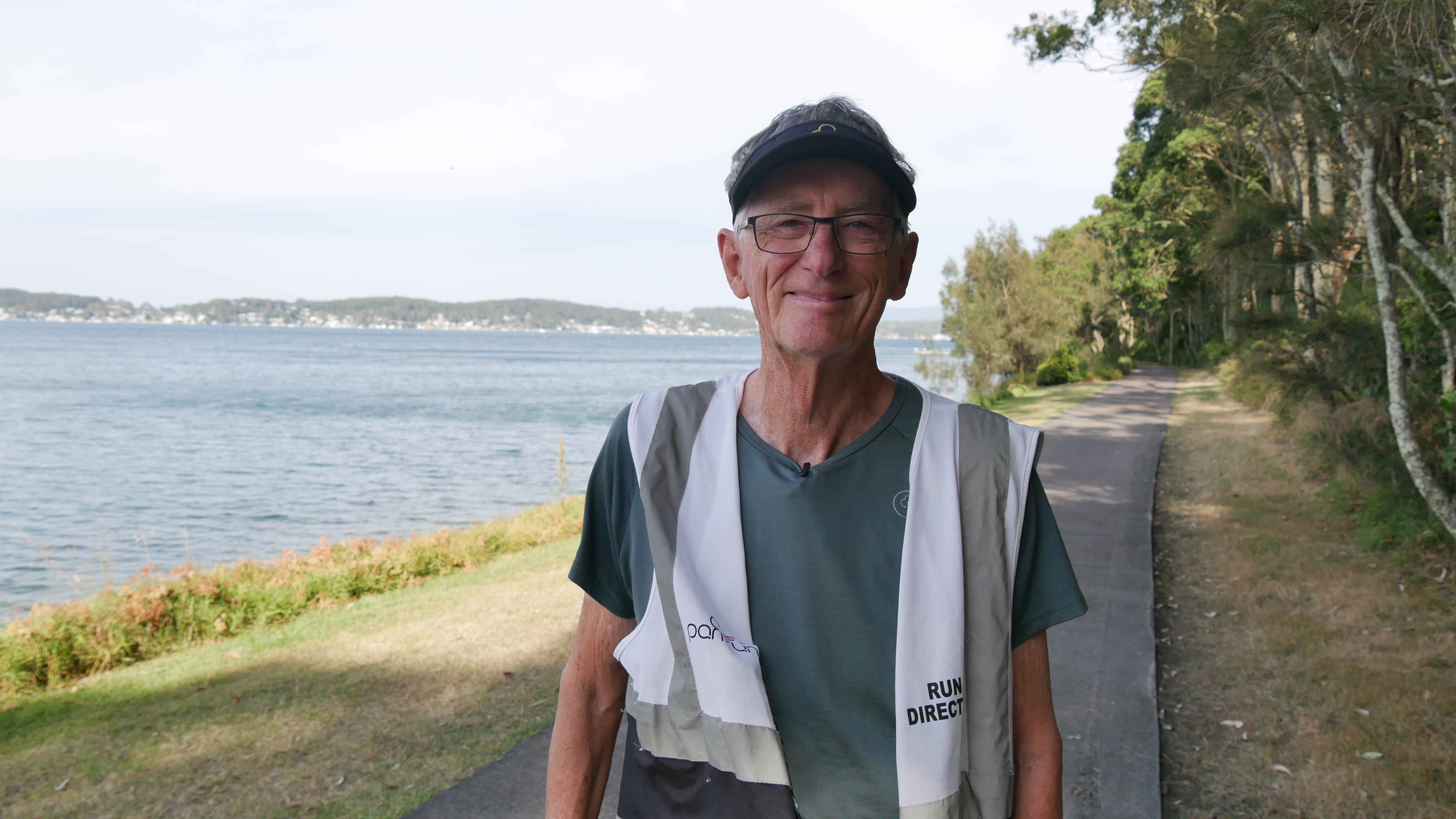 Peter Scotchmer de óculos, camisa de corrida, chapéu e colete de alta visibilidade em frente a uma trilha e um lago. 
