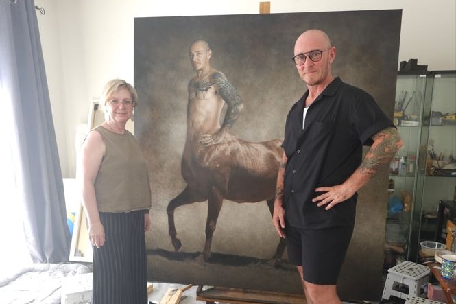 Jaq Grantford and Ed Le Brocq stand at either side of large canvas depicted Le Brocq as a centaur, in a cluttered studio.