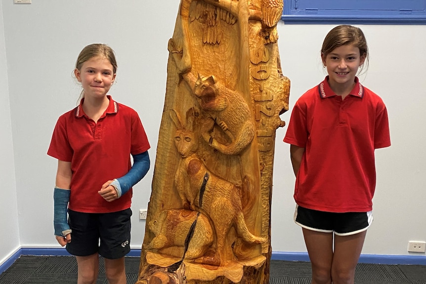 Two kids in a red t-shirt and black shorts stand next to a carved tree trunk. 