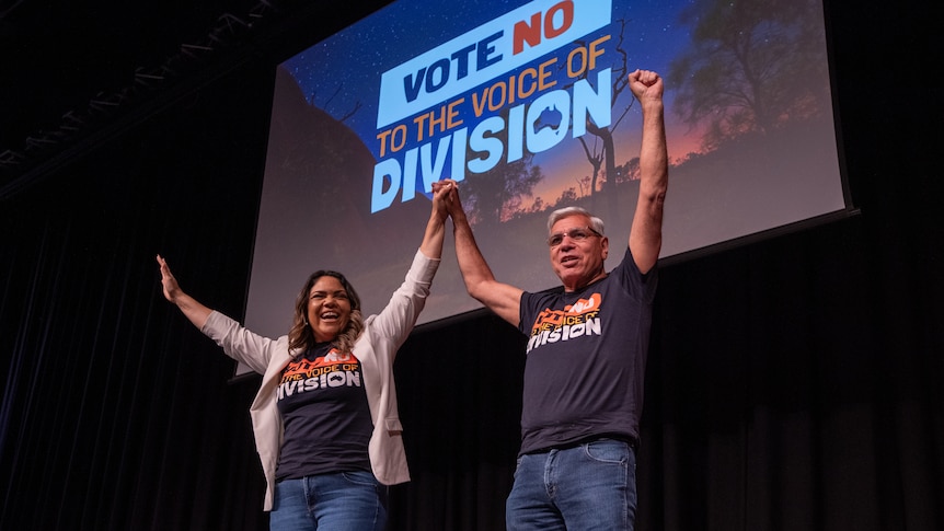 Two people sit on a stage smiling, both wearing t-shirts that say Vote No to the Voice of Division
