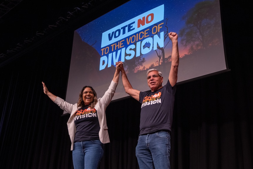 Two people sit on a stage smiling, both wearing t-shirts that say Vote No to the Voice of Division