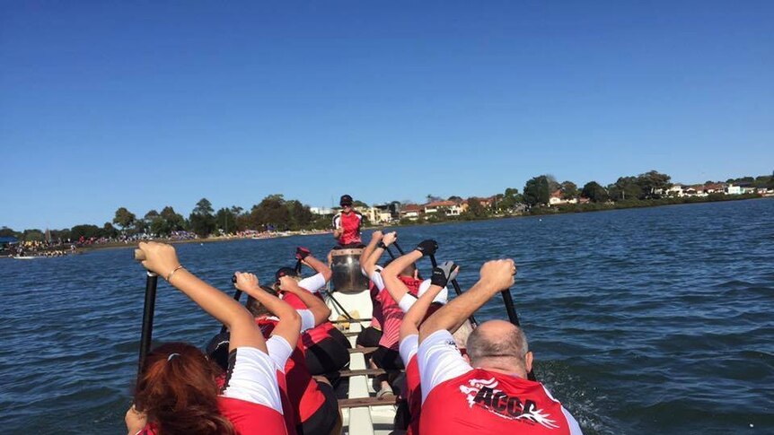 A tour group wearing red and white shirts rowing in a long boat on a NSW waterway.