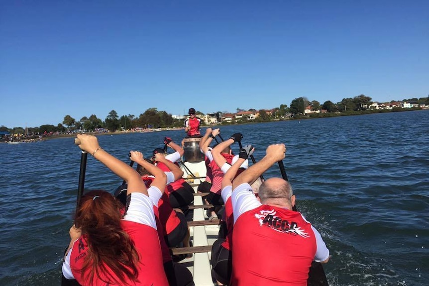 A tour group wearing red and white shirts rowing in a long boat on a NSW waterway.