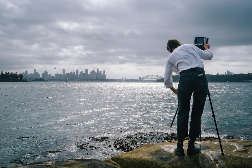 Sammy Hawker stands on the edge of Sydney Harbour balancing a large format camera on a tripod, the bridge visible in distance 