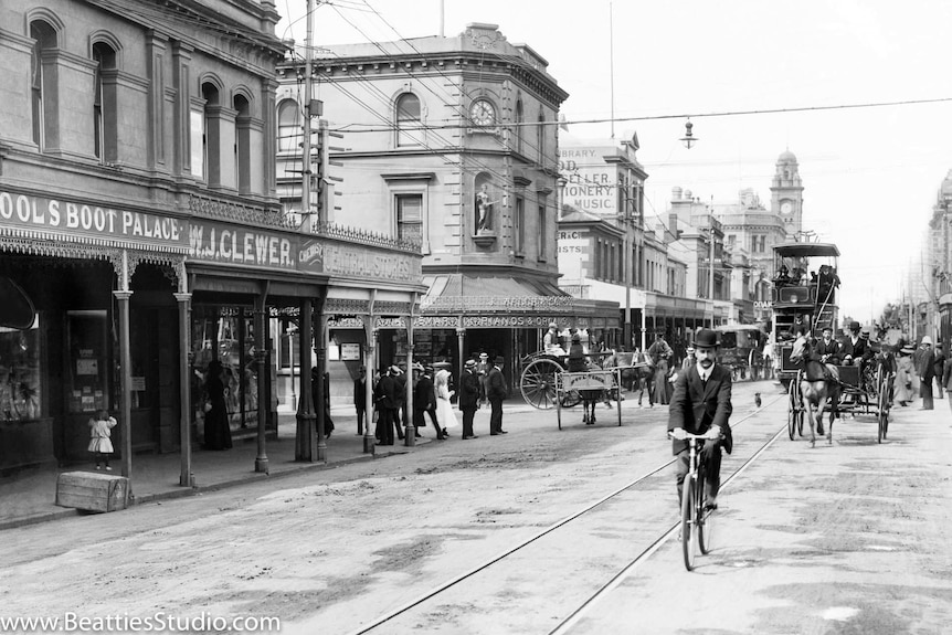 Black and white photo, man on bicycle in foreground, horse and cars, double decker trams and colonial era buildings