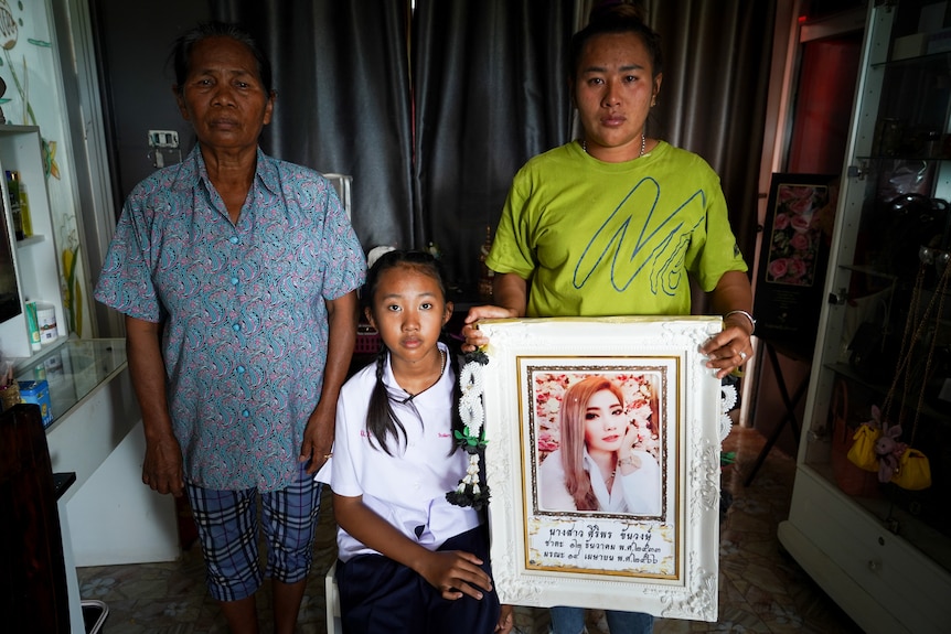 A girl sits on a chair next to her mother and her brother holding a portrait of her sister.