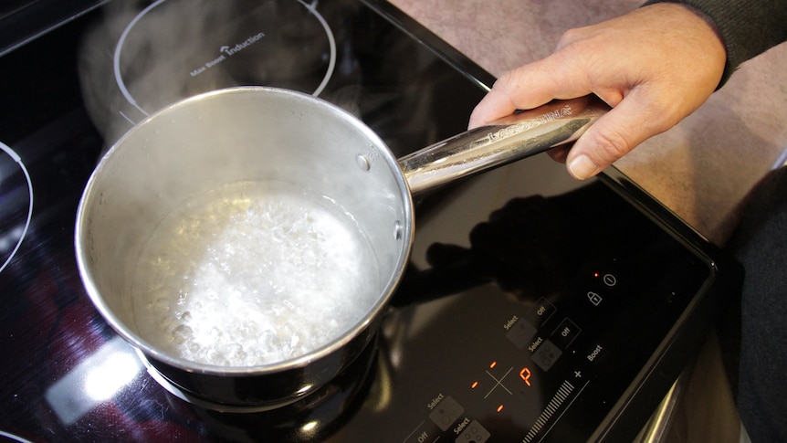 Someone holds the handle of a silver pot, boiling water on an induction stove top.