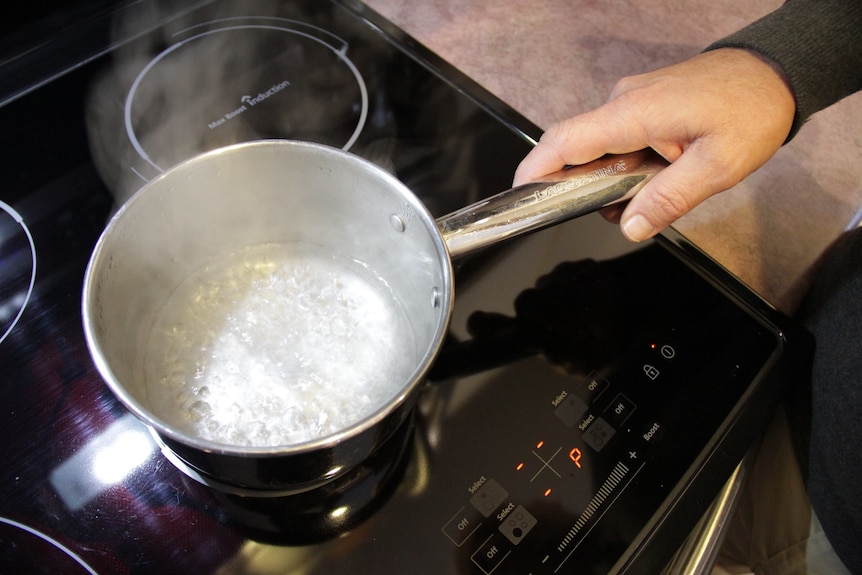 Someone holds the handle of a silver pot, boiling water on an induction stove top.