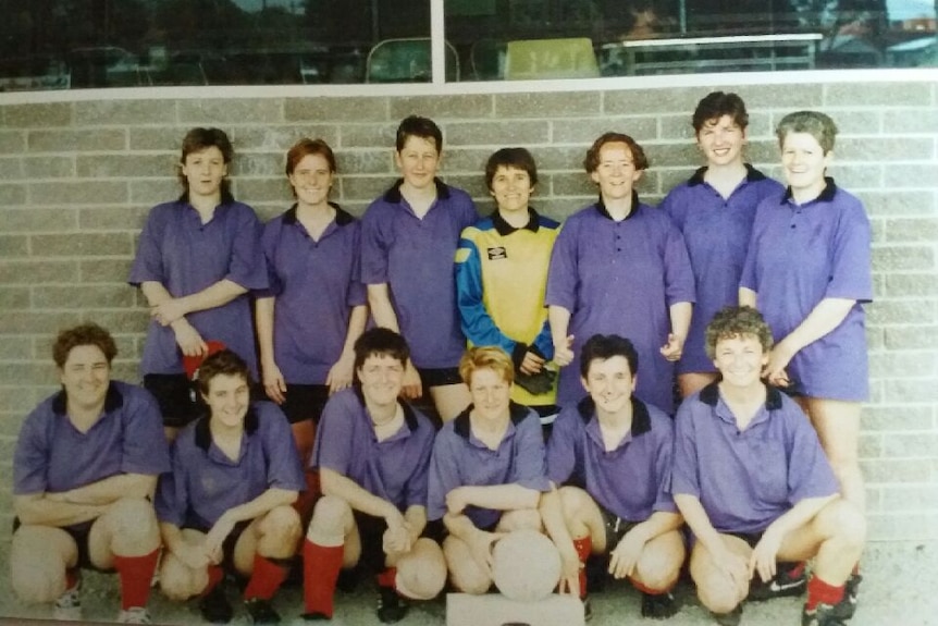 A group of female soccer players in purple shirts pose along a wall