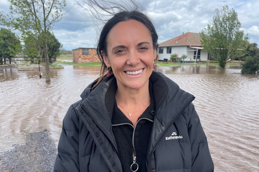 A smiling, dark-haired woman in a warm jacket stands in front of a flooded yard.