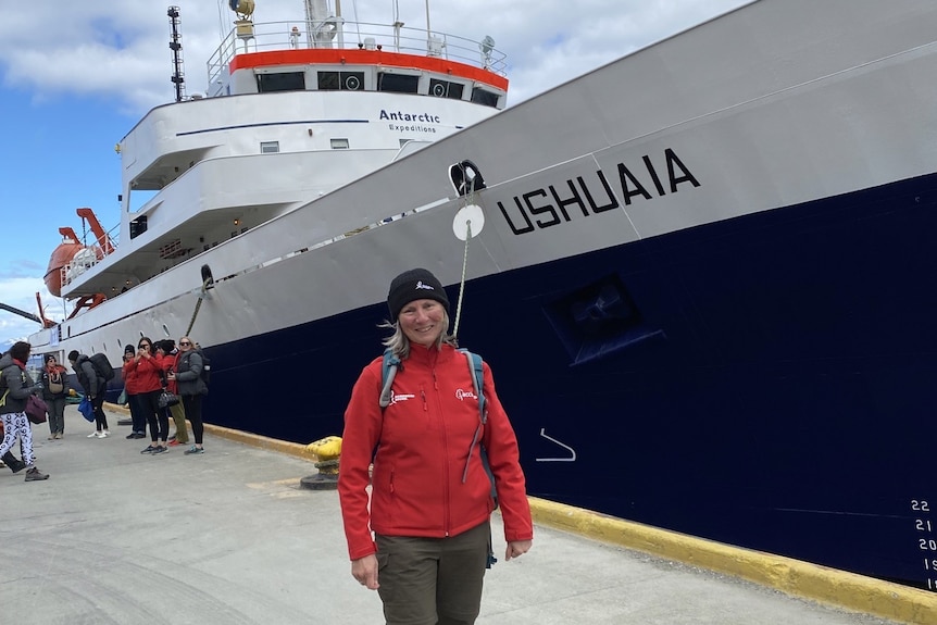 a woman in a red jacket stands in front of a large blue-hulled icebreaker