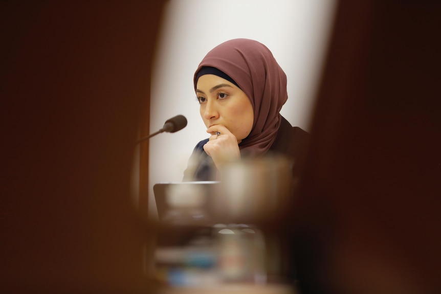 A young woman in a hijab listens as she sits behind a microphone.
