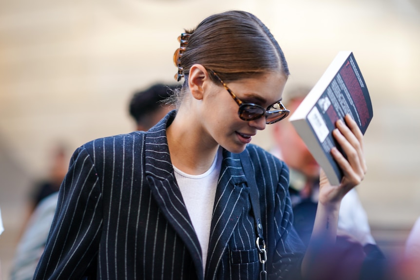 Kaia, in sunglasses, holds a black paperback up in front of her face as she walks.