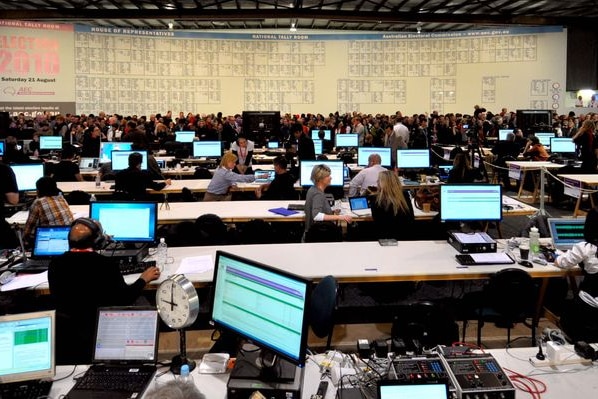 Journalists sit at their computers with members of the public and tally board in the distance