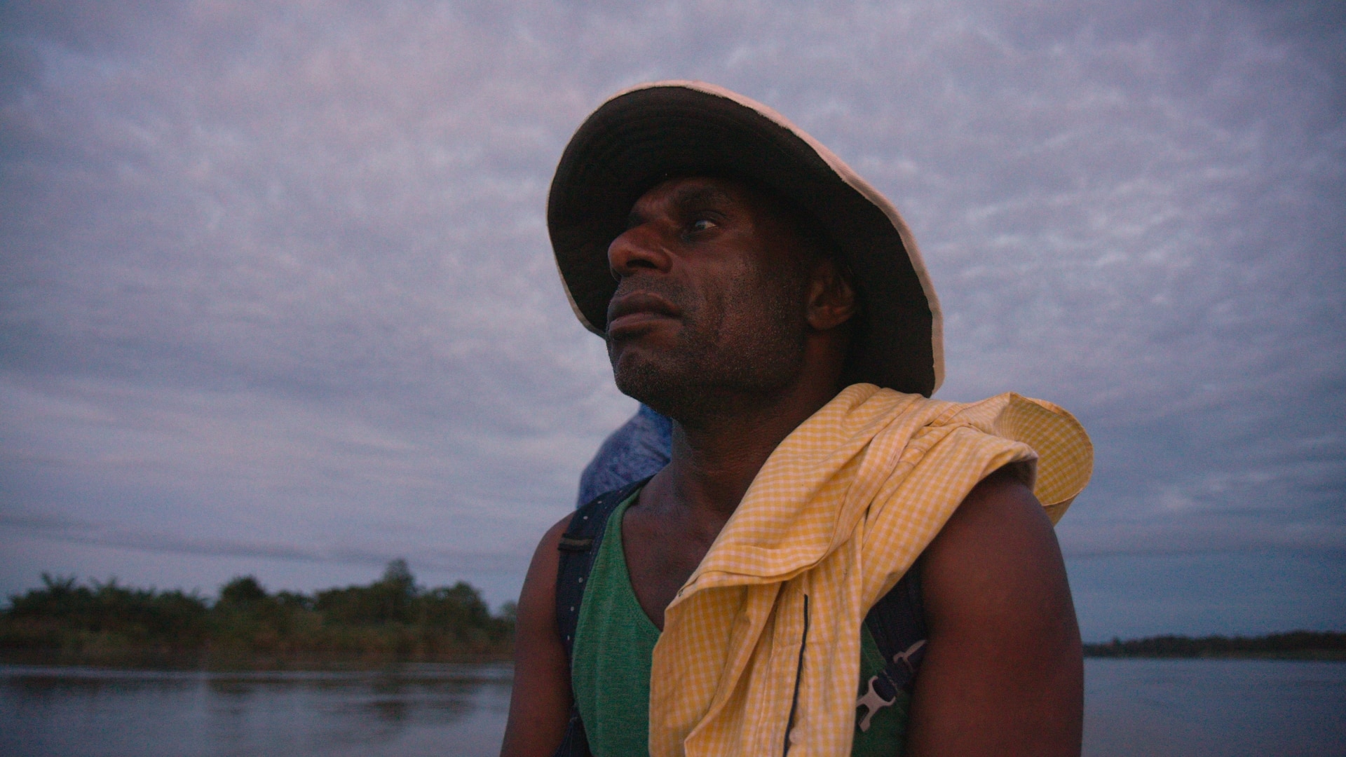 close up shot of a black man wearing a yellow brimmed hat looking at the river which courses behind him