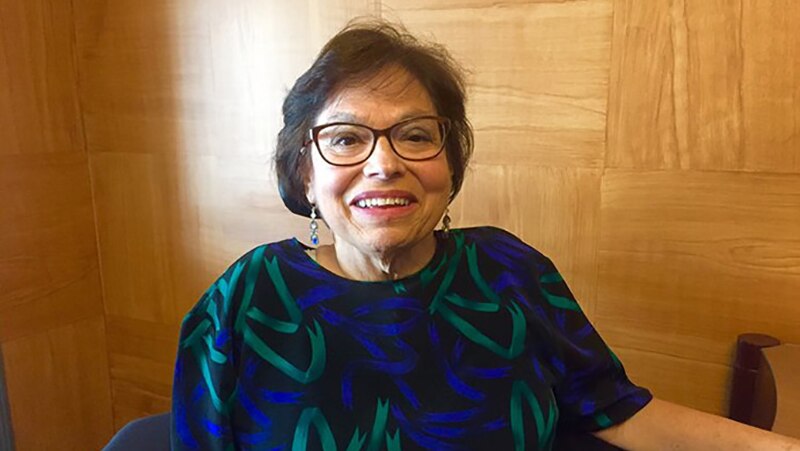 Photo of Judy  Heumann, a cis-gender white woman who is a wheelchair user with short brown hair & glasses. She is smiling kindly