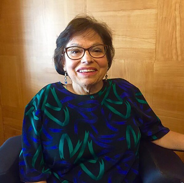 Photo of Judy  Heumann, a cis-gender white woman who is a wheelchair user with short brown hair & glasses. She is smiling kindly