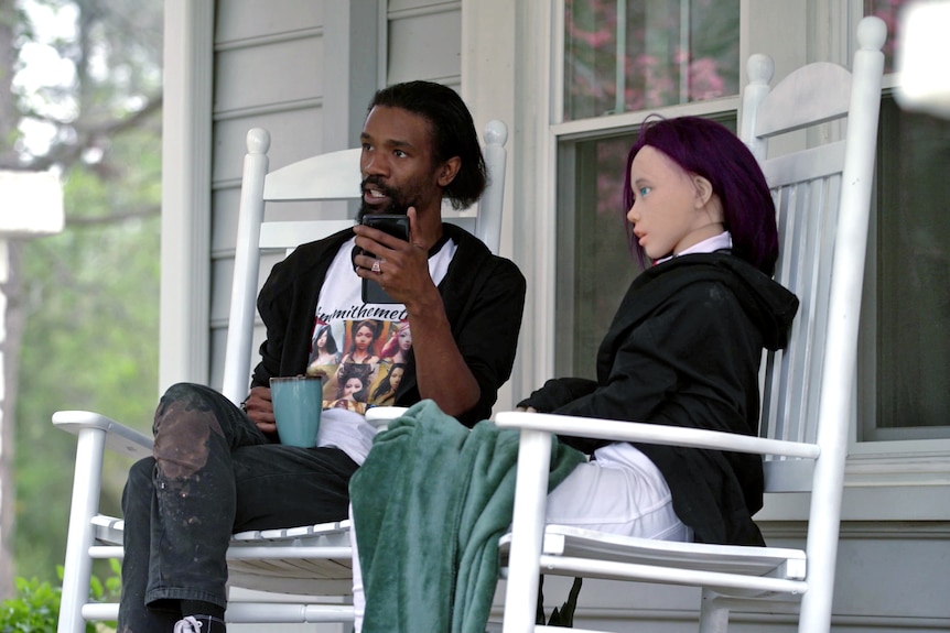 A man and a life-sized doll sit on a front porch. 