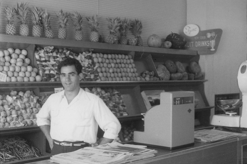 A black and white photo of a man standing behind a counter in a fruit and veggie store.