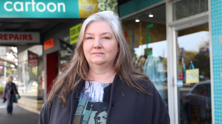 A woman with brown and white hair stands outside a shopping strip