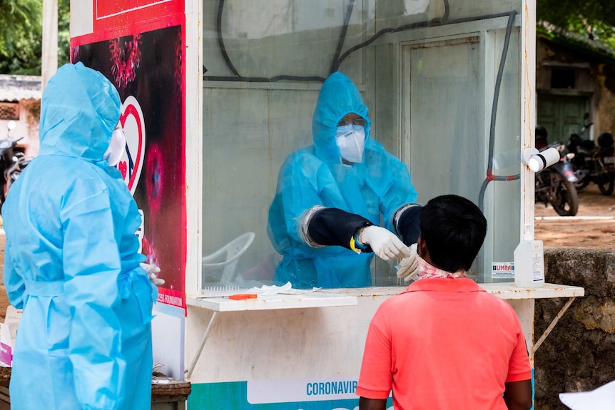 A health worker in full PPE testing someone for COVID-19 in a glass booth