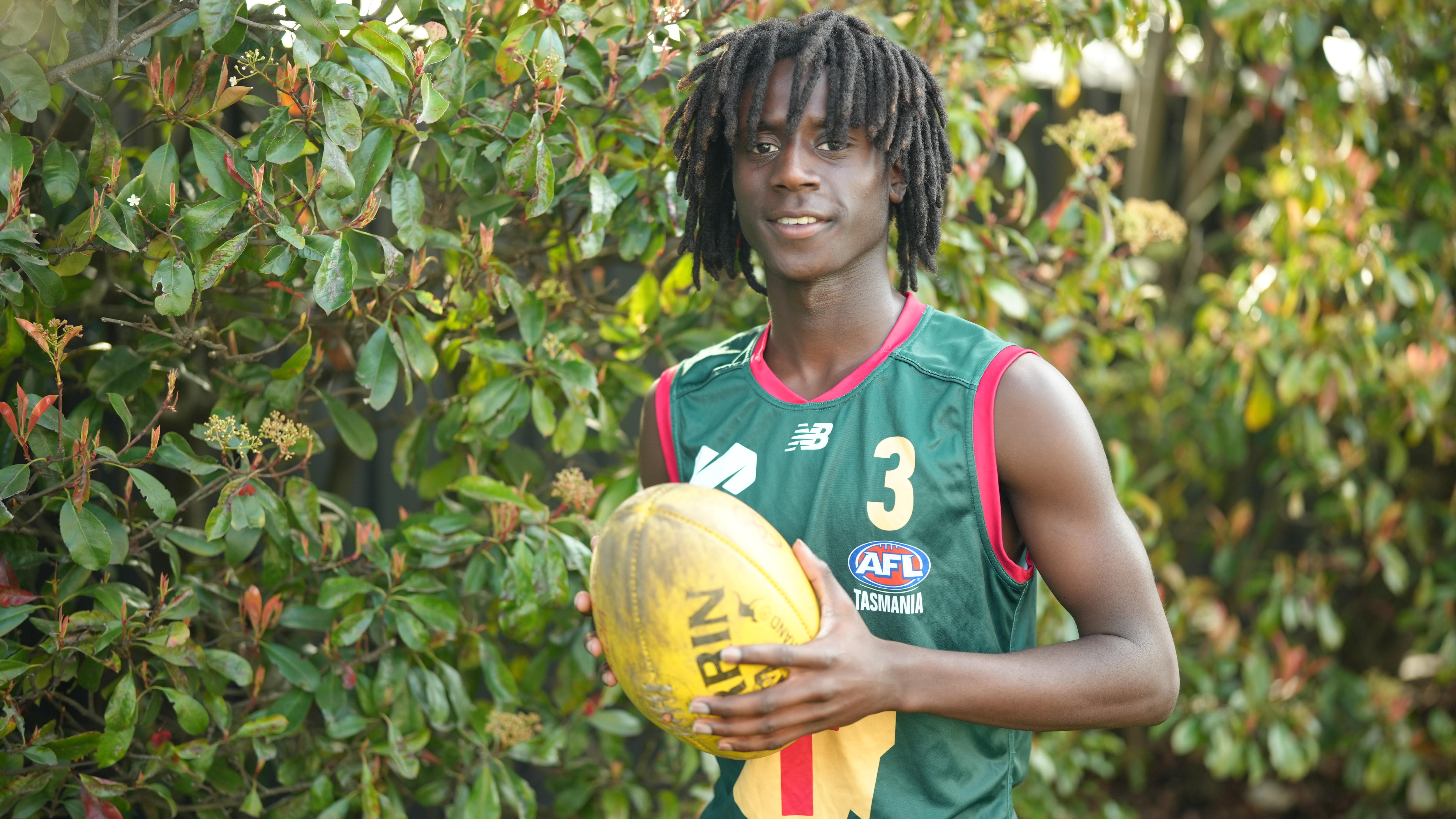 A young man wearing a green guernsey holding a yellow football and smiling.