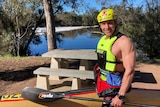 A smiling man in high-viz life jacket and helmet holds a kayak on a riverbank with trees and a picnic table.