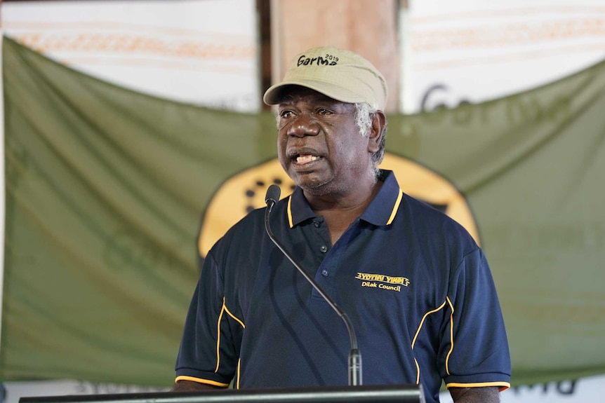 Djawa Yunupingu stands at a lecturn in front of the flag of the Gumatj people.