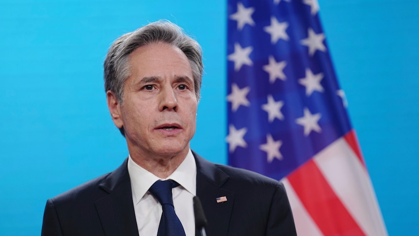 A man with grey hair in a black suit stands in front of the American flag.