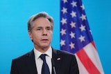 A man with grey hair in a black suit stands in front of the American flag.