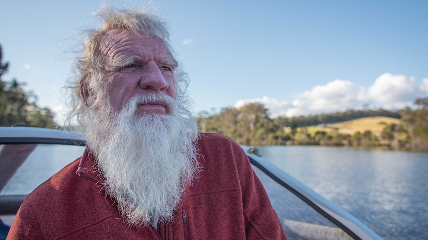 A man with white hair and a long white beard on a boat, the river and hills are behind as he stares into the distance.