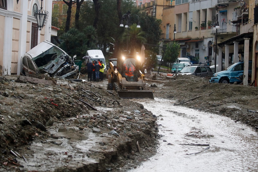 A truck pushes mud off the road as ruined buildings and cars line the street. 