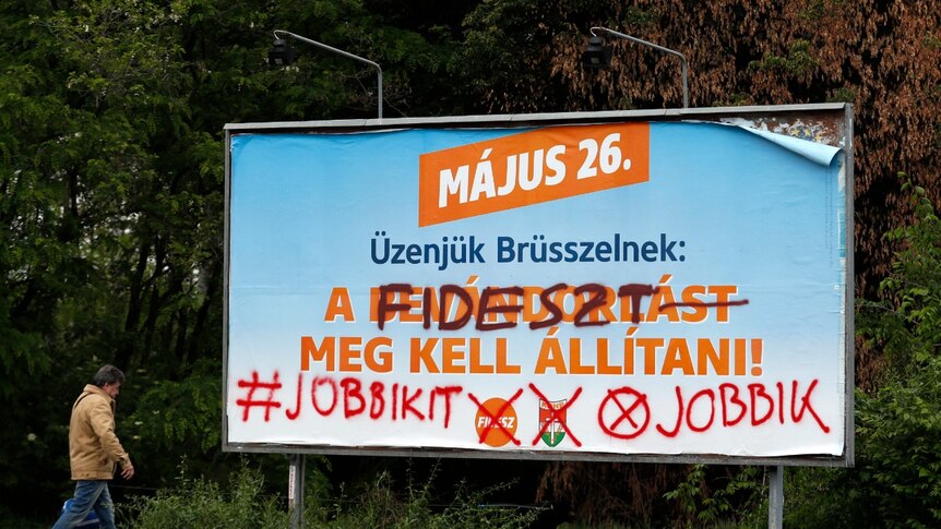A political campaign billboard in blue and orange is graffitied over with the name of its competitor.