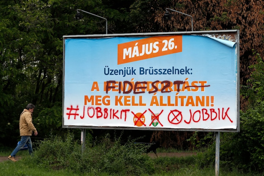 A political campaign billboard in blue and orange is graffitied over with the name of its competitor.