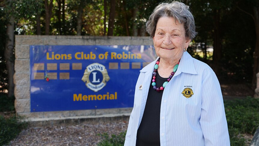 Elderly woman with a Lions club shirt on standing in front of the Lions Club of Robina memorial.