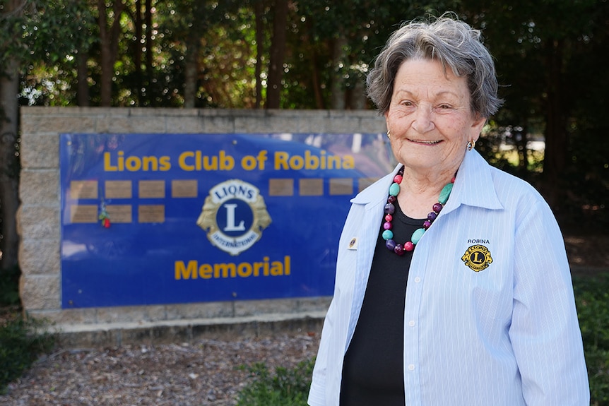 Elderly woman with a Lions club shirt on standing in front of the Lions Club of Robina memorial.