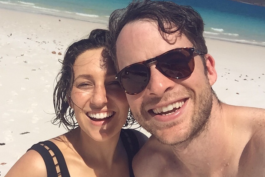 A couple smile at the camera on a beach in their swimwear