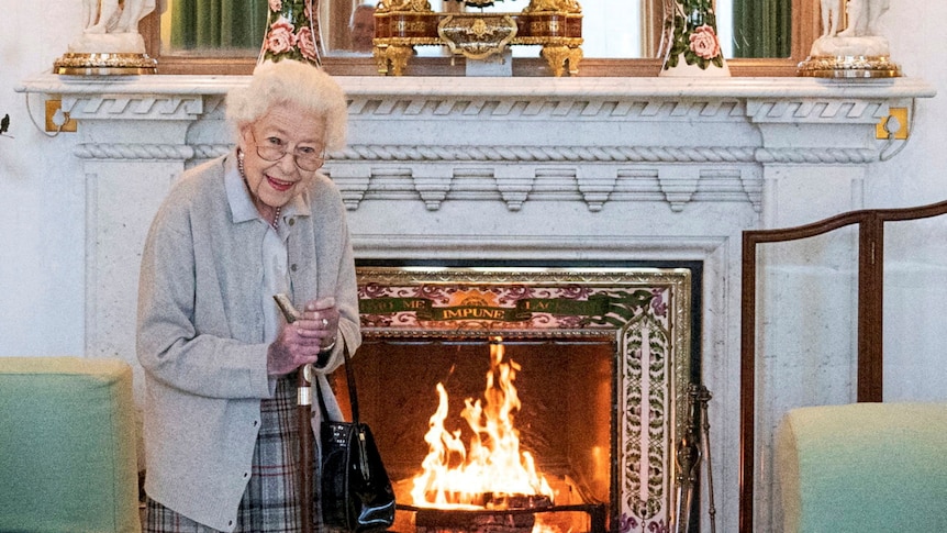 Queen Elizabeth stands smiling and leaning on a cane next to a lit fireplace.