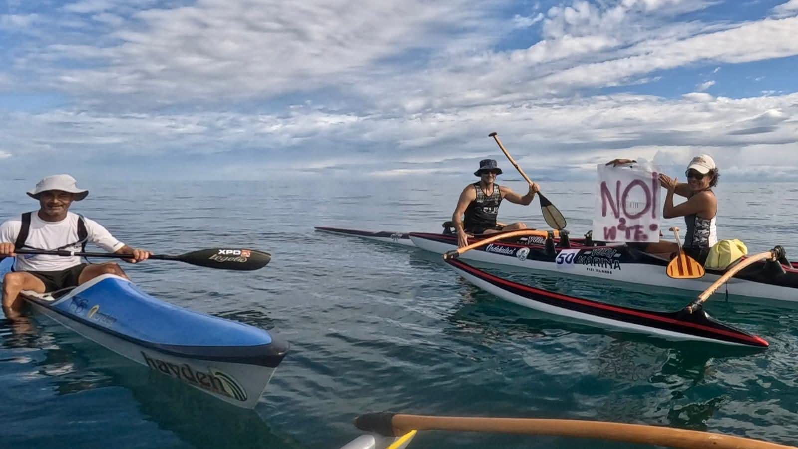 Two people are sitting in a canoe in open water, and a woman is holding a sign that says: "Not W2E"