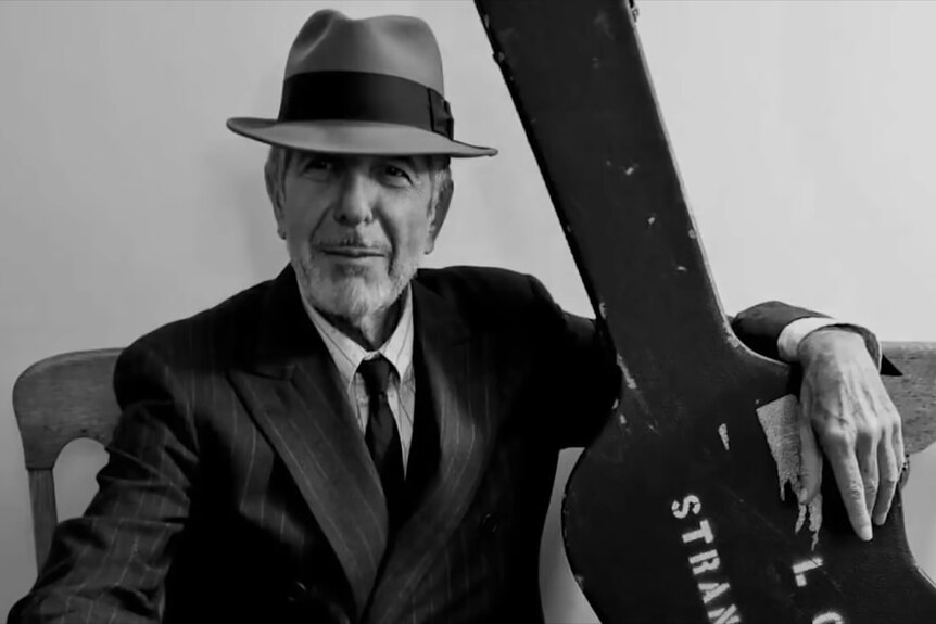 A B&W image of an older man wearing a suit and hat sitting with his arm around a guitar case, looking into the camera lens.