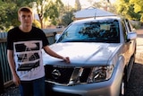 A young man stands next to a silver Nissan car. 