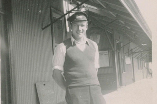 A black and white photo of a man wearing a hat, shirt and vest standing on a railway platform.
