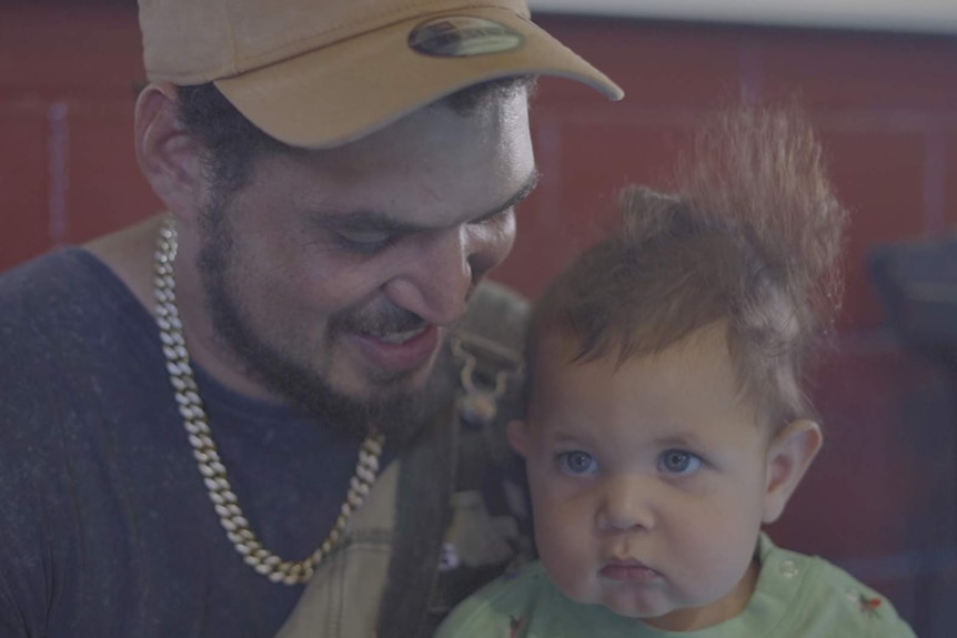 Wayne 'Dubb T' Tobane wearing a cap, grey shirt, smiling down at his young son who he's holding.