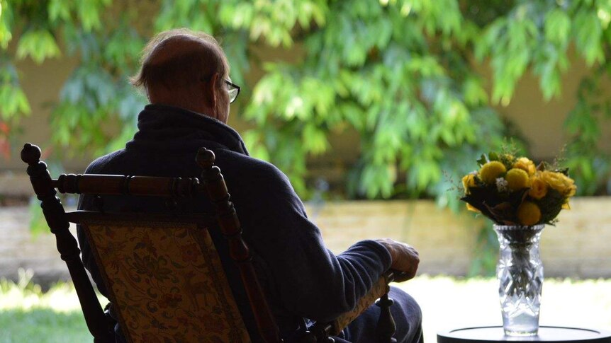 An elderly man sits in a rocking chair facing away into a sunlit garden, next to a vase of flowers on a table.