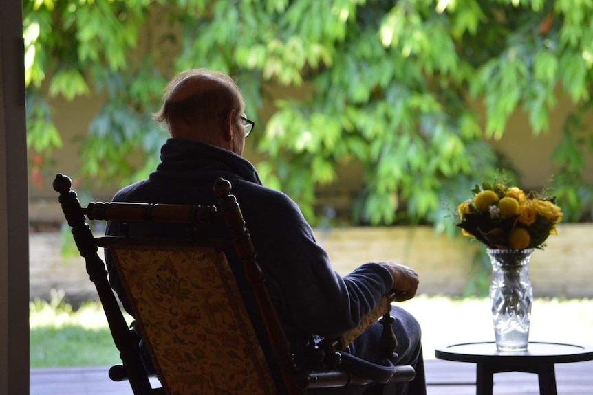 An elderly man sits in a rocking chair facing away into a sunlit garden, next to a vase of flowers on a table.