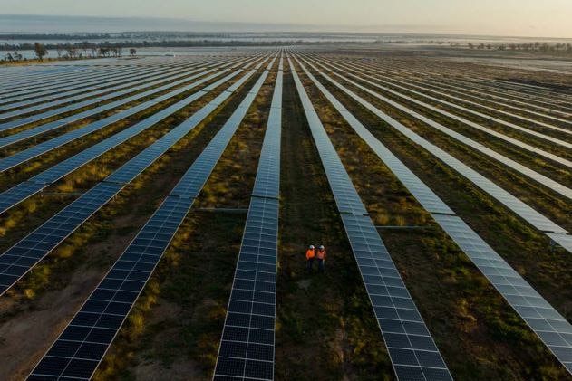 Due lavoratori in uniforme arancione camminano in una fila di pannelli solari in un enorme parco solare a Chinchilla.