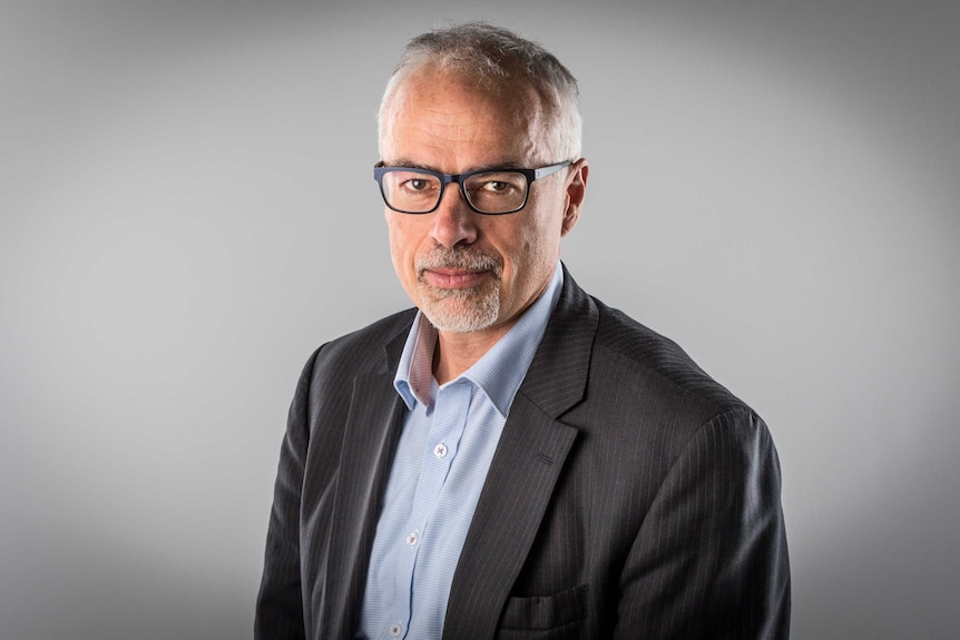 MCU of Professor Tony Blakely wearing black rimmed glasses, grey suit jacket and open-necked light blue shirt